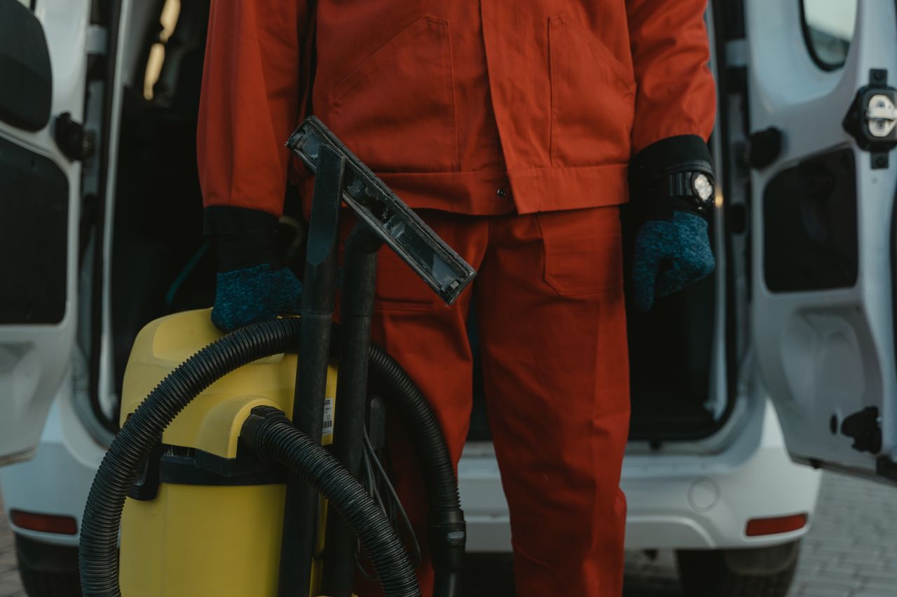 Un professionnel du nettoyage en uniforme rouge, aspirateur à la main près d’un van, prêt à intervenir.